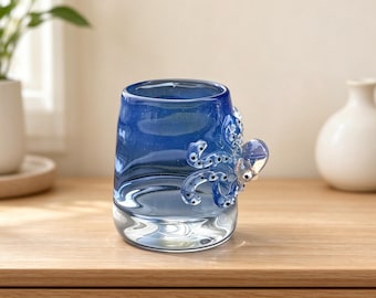 May include: A cylindrical glass mug featuring a deep blue gradient color and a three-dimensional octopus figure attached to the side. The octopus is light blue with dark blue suckers and small black eyes, integrated into the glass vessel design.