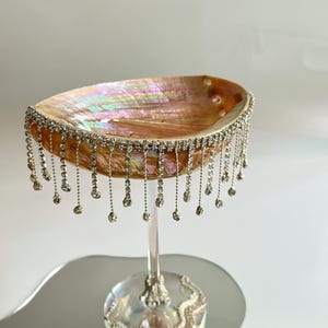 Peut inclure: Un gobelet décoratif doté d'une coupe en coquille d'ormeau irisée ornée d'une frange de strass transparents pendants. Le pied et la base circulaire sont en verre transparent, la base étant décorée d'un motif ondulé de petits strass.