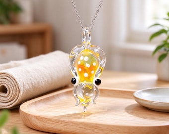 May include: A clear glass pendant shaped like a squid, featuring a yellow interior with white polka dots and two prominent black eyes. The pendant hangs from a thin silver-toned metal chain, showcasing a smooth, glossy finish throughout.