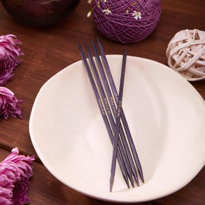 Könnte beinhalten: Ein Satz dunkelvioletter Stricknadeln auf einem cremefarbenen Teller. Die Nadeln, mit Text, werden von lila Garn und Blumen begleitet. Die Nadeln sind US-Größe 10, 6,00 mm.