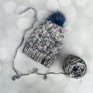 May include: A chunky knit beanie in shades of gray, white, and blue with a cable knit pattern and a dark blue faux fur pom-pom on top. A small black rectangular tag is attached to the brim. A matching ball of thick yarn is connected to the hat.