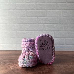 May include: Hand-knitted baby booties in pink, purple, and blue. One bootie is upright, the other shows the sole with the text "hello I'M NEW HERE" and an arrow. The booties are on a wooden surface.