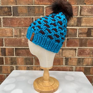 May include: A chunky knit beanie in turquoise blue with a brown geometric pattern and a brown faux fur pom-pom. The hat is displayed on a mannequin head. A small leather tag is sewn on the side.