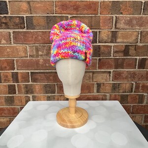 Op de afbeelding: Een levendige, handgebreide beanie in een regenboog van kleuren, waaronder roze, geel, oranje en blauw. De muts wordt getoond op een mannequinhoofd tegen een bakstenen muur. Een gezellig winteraccessoire.