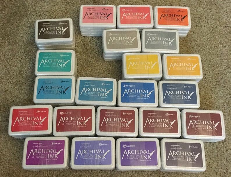 May include: A collection of 24 ink pads in various colors, all labeled "Ranger Archival Ink". The ink pads are arranged in a rectangular shape, with the top row having four ink pads, the second row having six ink pads, and the third row having six ink pads. The ink pads are all the same size and shape, and they are all made of a white plastic material.