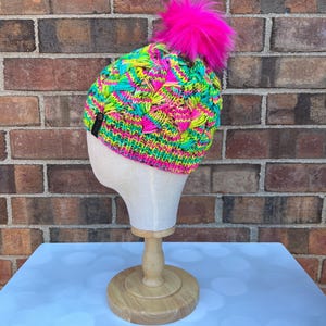 May include: A brightly colored knit hat with a vibrant mix of neon green, pink, and turquoise yarns. The hat features a textured pattern and is topped with a large, fluffy pink pom-pom. The hat is displayed on a mannequin head.