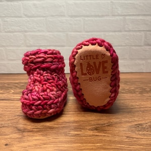 May include: A pair of handmade, pink and red crocheted baby booties. The sole of one bootie features the text "LITTLE LOVE BUG" in a heart-shaped design. The booties are made of a chunky yarn and sit on a wooden surface.