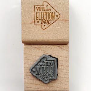 May include: Two wooden stamps. The top stamp has the words "VOTE ON ELECTION DAY" inside an arrow-shaped design. The bottom stamp shows the reverse image of the text and design. The stamps are made of light-colored wood.