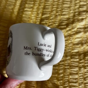 May include: A white ceramic mug featuring a curved handle and black printed text that reads: Lucie and Mrs. Tiggy-winkle with the bundles of clothes. The mug has a smooth, glossy finish and a cylindrical shape with a visible illustration on the side.