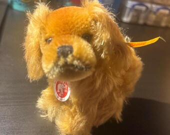 May include: A plush golden-brown dog toy with long, soft fur, dark eyes, and a black nose. A small white circular tag with a red elephant graphic and the text "Steiff" is attached to the chest, and a yellow ribbon tag extends from the side.