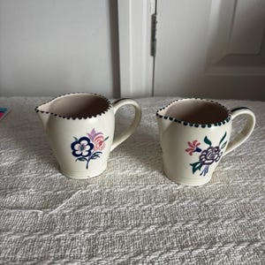 Peut inclure: Deux pichets en céramique de couleur crème dotés d'un bord festonné avec une bordure à pois vert foncé. Chaque pichet possède une anse et un motif floral peint à la main dans des tons de bleu, rose, violet et vert sur la surface avant.
