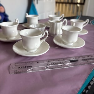 Puede incluir: Un juego de tazas de té y platillos en miniatura de cerámica blanca, una pequeña jarra de leche y un cuenco. Una regla de plástico transparente está colocada en primer plano, mostrando la escala de los objetos en centímetros, hasta 15 cm.