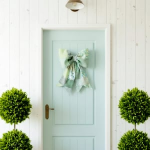 May include: A large decorative bow made from a patchwork of various light green, sage, and white floral patterned fabrics. The bow features two wide loops and two long, pointed tails, and it is mounted centrally on a light blue paneled door.