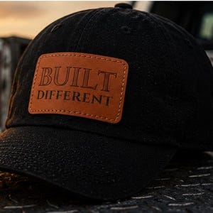May include: A black baseball cap featuring a rectangular tan leather patch centred on the front. The patch is embossed with the text BUILT DIFFERENT in a serif font. The cap has a curved brim and a structured crown with visible stitching.