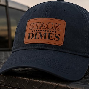 May include: A navy blue baseball cap featuring a rectangular tan leather patch on the front. The patch is embossed with the text STACK DIMES in a bold, dark font, separated by a horizontal row of six small dots between the two words.