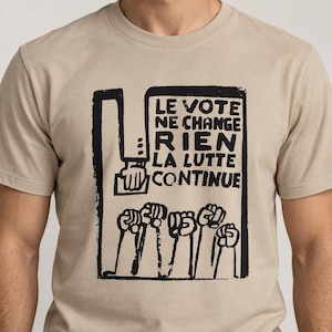May include: A beige crew neck t-shirt featuring a black graphic print of a hand placing a ballot into a box above a row of raised fists. The text reads: LE VOTE NE CHANGE RIEN LA LUTTE CONTINUE. The design has a bold, hand-drawn aesthetic.