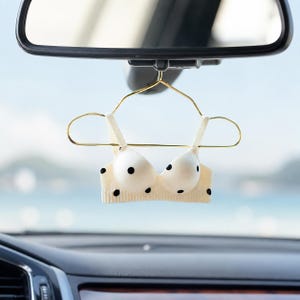 May include: A miniature cream-colored bra with black polka dots hanging from a gold-toned wire hanger. The small garment features molded cups and a ribbed band, suspended from the rearview mirror of a vehicle against a blurred outdoor backdrop.