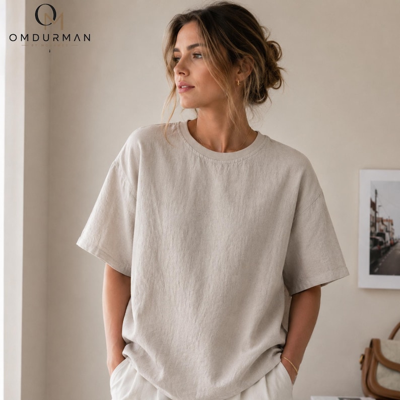 May include: A beige short-sleeved t-shirt with a round neckline and a loose, relaxed fit. The fabric has a visible textured weave. The top left corner features the black text OMDURMAN. The garment is worn in a casual, untucked style.