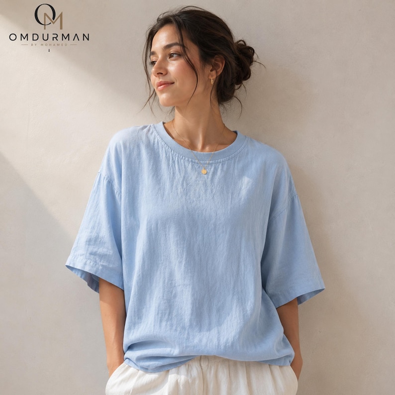 May include: A light blue short-sleeved t-shirt with a crew neckline and a relaxed, loose-fitting silhouette. The shirt features a solid color design and is paired with white bottoms. The OMDURMAN BY MOHAMED logo is visible in the top left corner.