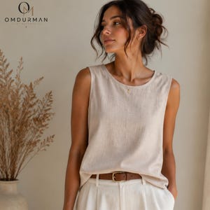 Puede incluir: Una blusa sin mangas de lino beige con cuello redondo, combinada con pantalones blancos y un cinturón de cuero marrón con hebilla dorada. La prenda tiene una textura natural y un corte relajado, acompañada por un collar de cadena dorada.