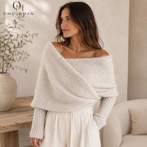 Könnte beinhalten: Ein schulterfreier beiger Strickpullover mit einem gewickelten Oberteil in strukturierter Optik und langen gerippten Ärmeln. Das Kleidungsstück wird mit einer hellen Hose kombiniert. Das OMDURMAN-Logo ist oben links zu sehen.