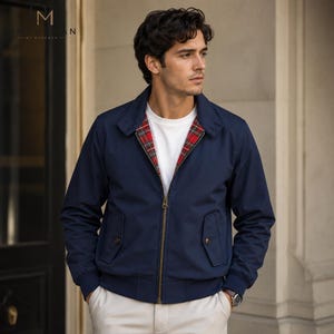 May include: A navy blue zip-up jacket featuring a red, green, and yellow plaid lining, a pointed collar, and two front flap pockets with buttons. The jacket is worn over a plain white crew neck t-shirt and paired with light-colored trousers.