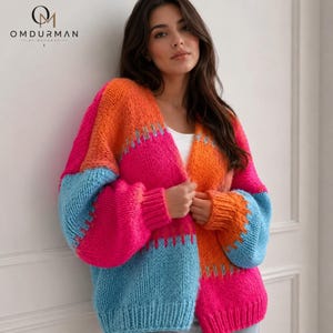 Könnte beinhalten: Eine grob gestrickte Strickjacke mit einem lebendigen Color-Block-Design in leuchtendem Pink, Orange und Hellblau. Der offen zu tragende Pullover hat lange Ärmel mit gerippten Bündchen und Saum sowie eine dicke, texturierte Garnstruktur.
