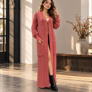 Könnte beinhalten: Eine lange, vorne offene Strickjacke mit Zopfmuster in Altrosa. Das bodenlange Kleidungsstück verfügt über lange Ärmel mit gerippten Bündchen, einen tiefen V-Ausschnitt und zwei große aufgesetzte Taschen auf der Vorderseite des Strickmusters.