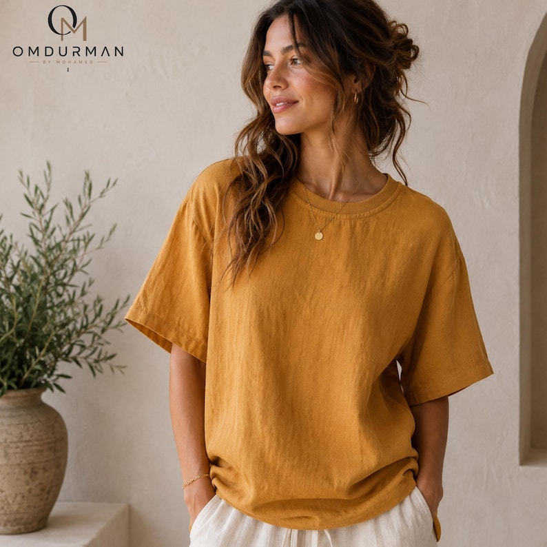 May include: A mustard yellow short-sleeved t-shirt with a crew neckline and a relaxed fit. The shirt features a visible textured fabric weave. The OMDURMAN BY MOHAMED logo is printed in the upper left corner of the image in black text.
