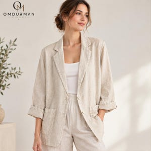 Puede incluir: Un blazer de color beige con solapa de muesca, dos botones frontales y dos bolsillos de parche. El blazer presenta mangas de tres cuartos con puños doblados y se lleva sobre una prenda superior blanca. El logo OMDURMAN BY MOHAMED es visible arriba a la izquierda.