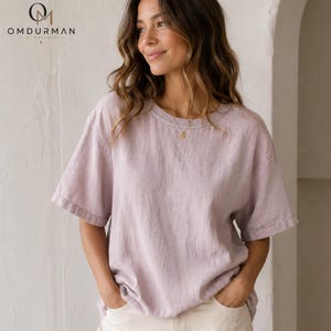 May include: A light lavender short-sleeved t-shirt with a crew neckline and a relaxed fit. The fabric has a visible textured weave. A small gold circular pendant necklace rests at the base of the neck. The logo OMDURMAN BY MOHAMED is visible.