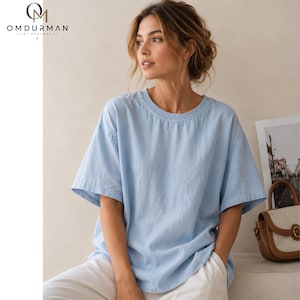 May include: A light blue short-sleeved t-shirt with a crew neckline and a relaxed fit. The shirt features a solid color design with visible fabric texture. The OMDURMAN BY MOHAMED logo is printed in the upper left corner of the image.