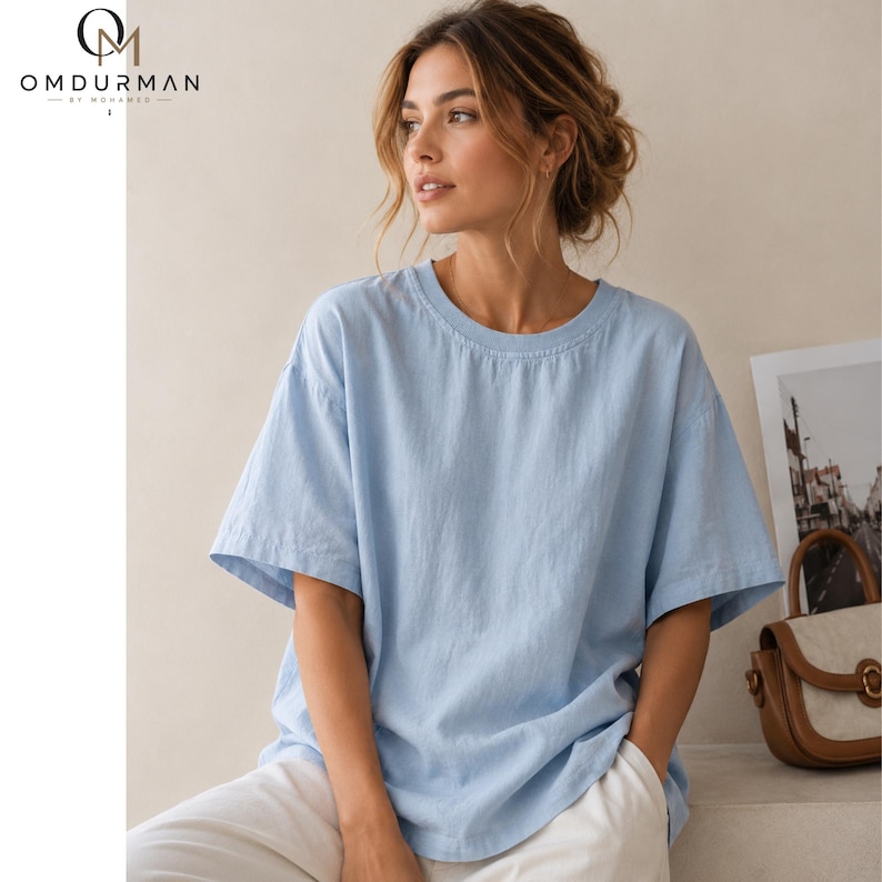 May include: A light blue short-sleeved t-shirt with a crew neckline and a relaxed fit. The shirt features a solid color design with visible fabric texture. The OMDURMAN BY MOHAMED logo is printed in the upper left corner of the image.