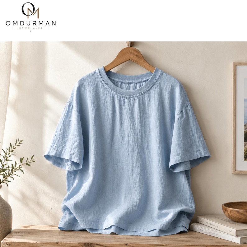 May include: A light blue short-sleeved t-shirt with a round neckline, displayed on a wooden hanger. The shirt features a textured fabric surface and a loose, relaxed fit. The brand logo OMDURMAN BY MOHAMED is visible in the top left corner.