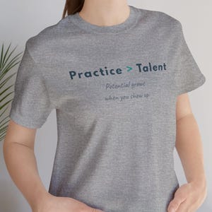 Puede incluir: Una camiseta gris jaspeado de manga corta con el texto azul oscuro Practice > Talent impreso en el pecho. Debajo, un texto más pequeño en azul oscuro dice Potential grows when you show up. La camiseta tiene un cuello redondo estándar.