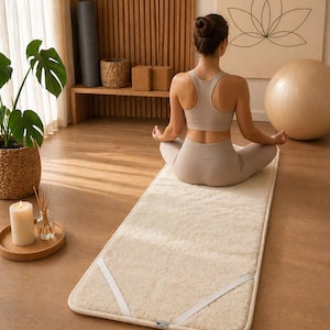 May include: A rectangular, off-white, plush fleece yoga mat featuring rounded corners and two white elastic corner straps. The mat has a thick, textured, wool-like surface and a finished border, designed for floor-based exercise and stretching.