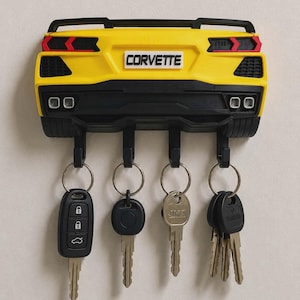 May include: A yellow and black wall-mounted key holder shaped like the rear of a Corvette sports car. The unit features a black spoiler, red taillights, and a white number plate with the text CORVETTE. Four black hooks hold various metal keys.