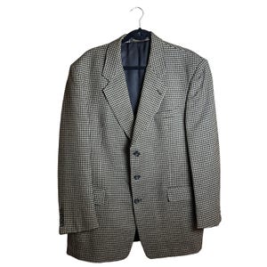 Könnte beinhalten: Ein Blazer mit schwarz-weißem Hahnentrittmuster. Das Kleidungsstück verfügt über ein steigendes Revers, drei dunkle Knöpfe auf der Vorderseite, zwei Pattentaschen in der Taille und lange Ärmel mit kleinen Knöpfen an den Manschetten. Innen dunkel gefüttert.