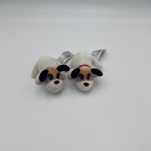 May include: Two small plush dog toys featuring white bodies, tan patches around the eyes, and black ears. Each toy has a red collar and a black nose, with a white fabric tag extending from the back of the head, designed for soft play.