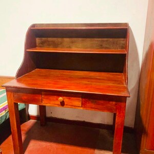Könnte beinhalten: Ein rotbrauner Schreibtisch aus Holz mit einer flachen Arbeitsfläche, einer einzelnen mittigen Schublade mit einem kleinen runden Knopf und einem aufgesetzten Regal mit zwei offenen Fächern. Der Tisch steht auf vier geraden Beinen.