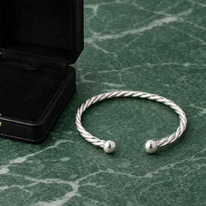 Puede incluir: Una pulsera tipo brazalete de color plateado con una textura trenzada similar a una cuerda en toda la banda. La pulsera es abierta y termina con una cuenta esférica lisa y pulida en cada extremo, presentada junto a un joyero negro abierto.