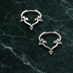 Könnte beinhalten: Ein Paar silberne Creolen mit einem herzförmigen Design, das an der Unterseite und an den Seiten mit kleinen Perlen verziert ist. Die Ohrringe verfügen über einen Klappverschluss und eine polierte Metalloberfläche mit detaillierten Verzierungen.