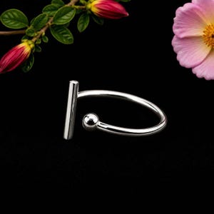 Può includere: Un anello aperto in metallo argentato lucido con una barra verticale su un'estremità e una piccola perla sferica sull'altra. La fascia è liscia e circolare, creando una finitura metallica pulita e riflettente contro lo sfondo scuro.
