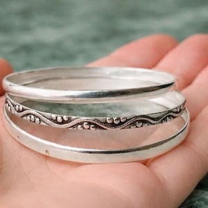 May include: A set of three silver-toned bangles. The top and bottom bangles feature a smooth, polished surface, while the middle bangle displays a textured, wavy pattern with small raised dots along the entire circumference of the metal band.