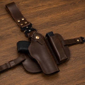 May include: A dark brown leather holster system featuring a moulded firearm pouch and an integrated magazine carrier. The assembly includes adjustable straps with black metal quick-release buckles and brass-toned snap fasteners for secure attachment.