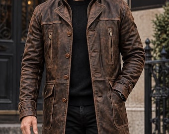 May include: A distressed brown leather trench coat featuring a high collar, vertical zippered chest pockets, and two large flap pockets at the waist. The coat has button closures down the front and adjustable cuffs with button tabs, worn over a black sweater.