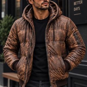 May include: A brown distressed leather hooded puffer jacket with a front zipper closure, ribbed cuffs, and a zippered utility pocket on the left sleeve. The jacket is worn over a black shirt and dark gray jeans, paired with black sunglasses.