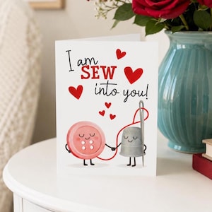 May include: A white greeting card featuring a cartoon pink button and a grey thimble holding hands with a red thread. The text reads I am SEW into you! with several red hearts scattered around the illustration on the front of the card.
