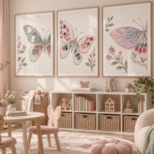 Könnte beinhalten: Drei gerahmte Wandbilder mit detaillierten Schmetterlingsillustrationen und floralen Akzenten in sanften Pastelltönen. Die Kunstwerke zeigen komplexe Flügelmuster in Rosa-, Blau- und Grüntönen und hängen über einem weißen Regalmöbel.