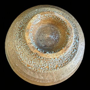May include: A circular ceramic bowl viewed from the bottom, featuring a textured, sandy-brown exterior surface with a raised foot ring. The base displays a mottled, light-green glaze pattern that fills the crevices of the rough, porous clay texture.
